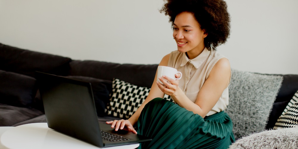 A woman sitting on a couch using a laptop while drinking a coffee