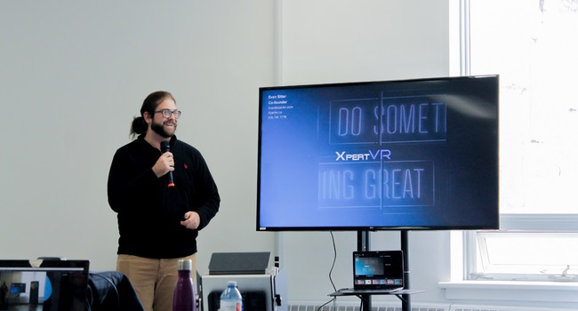 A man holding a microphone giving a presentation next to a television screen