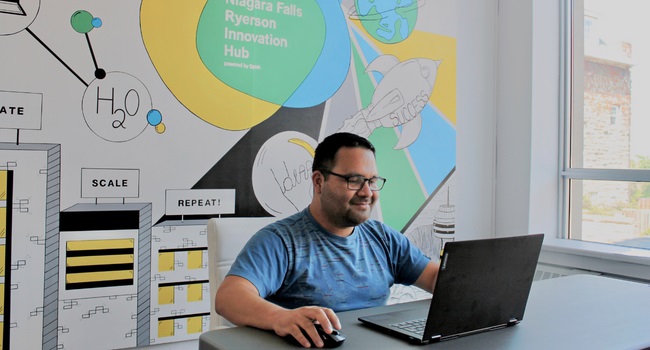 A man sitting at a desk in front of a laptop with a mural behind him
