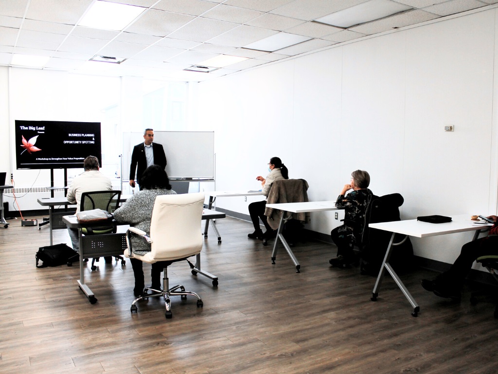 A man giving a presentation in a large open room while several people sit at tables and watch