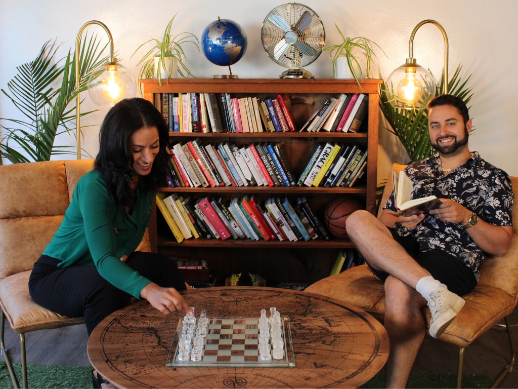 A man holding a book and smiling while a woman seated beside him moves a chess piece