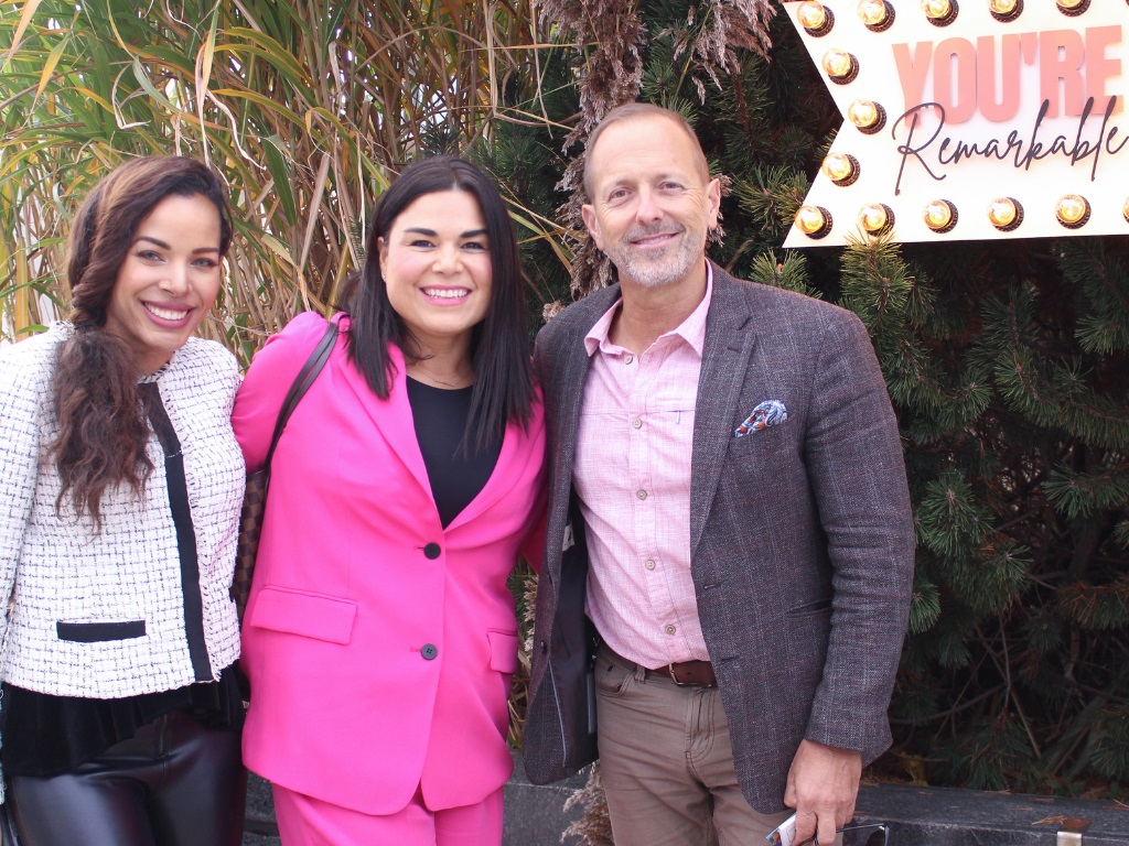 Two women and a man pose for a photo behind a sign saying: Youre Remarkable