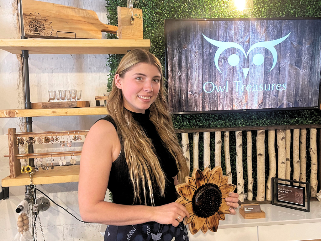 Woman standing in front of a wooden shelf display at the Magnificent Women's Market. The sign behind the woman reads "Owl Treasures," the name of her business.